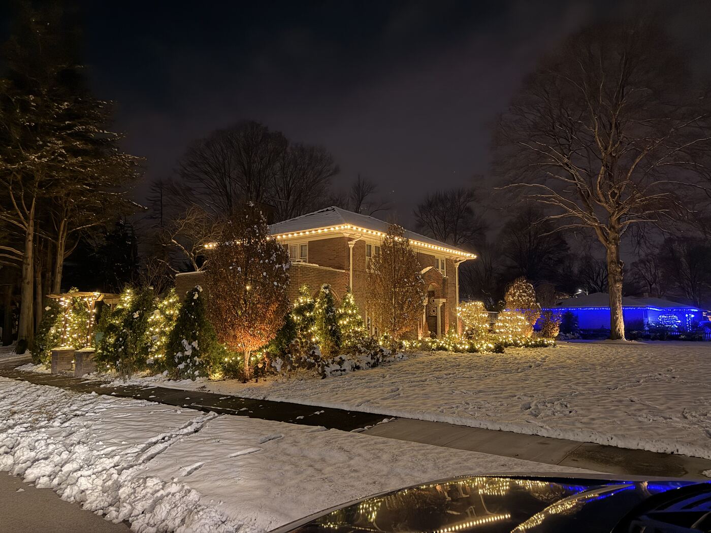 Warm white holiday lighting on a brick home's roofline with wrapped shrubs and trees in the snow